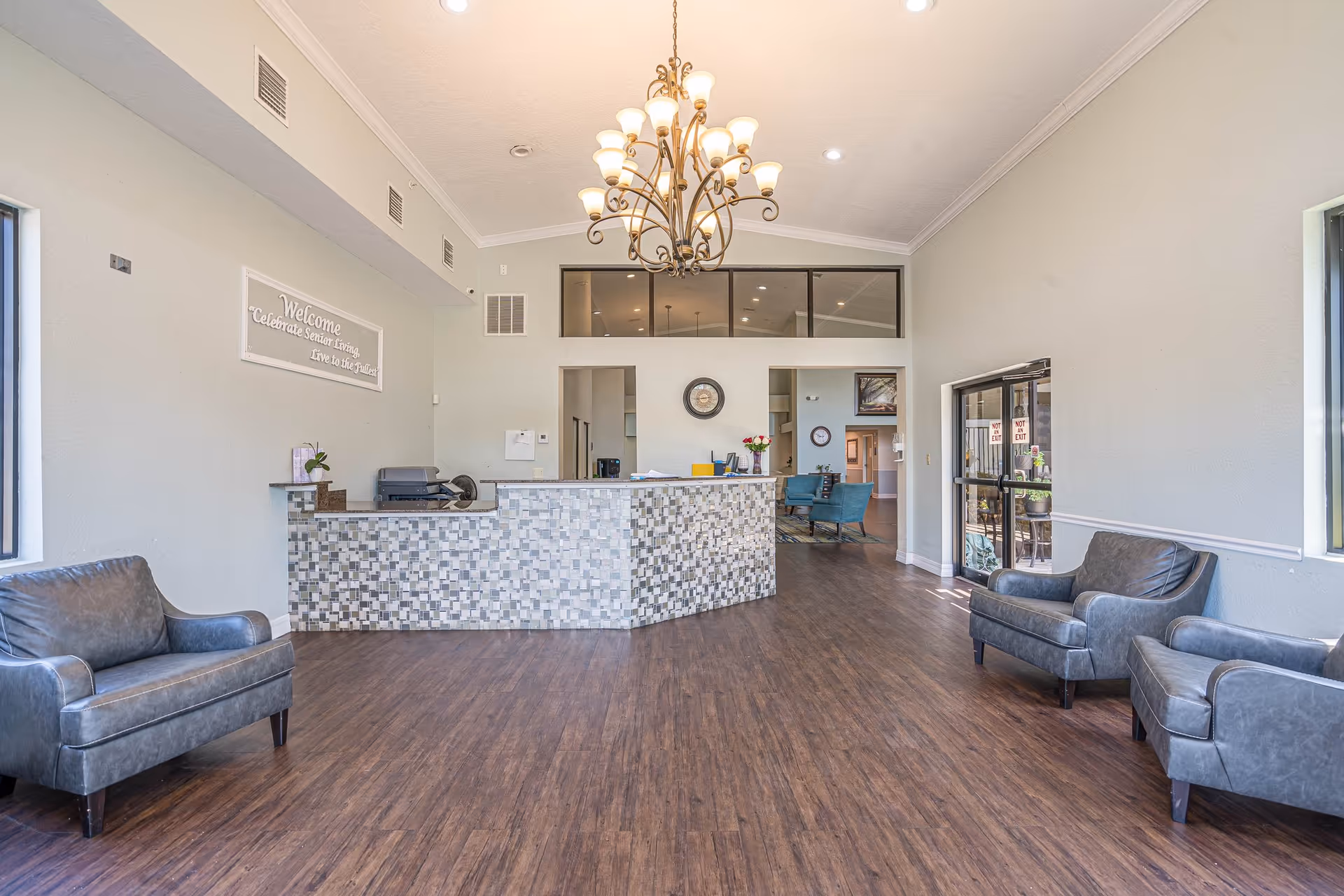 Spacious reception area with a tiled front desk, three gray leather armchairs, wooden flooring, large windows, and a chandelier hanging from the ceiling. A sign on the wall reads 'Welcome Celebrate Senior Living Live to the fullest.'
