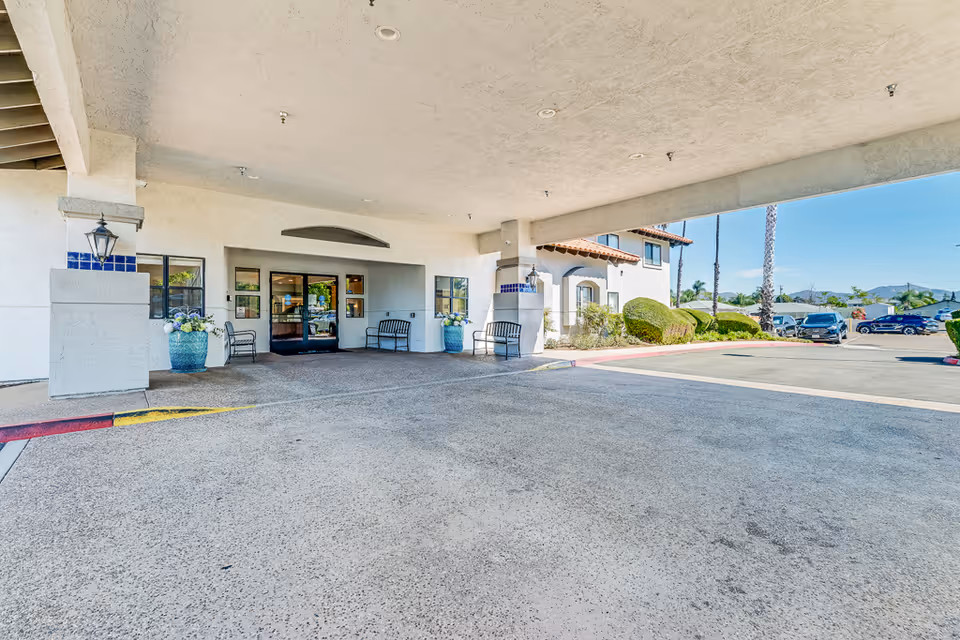 Covered entrance area of a senior living facility with benches, large planters with flowers, and glass doors leading inside. The building exterior is white with blue tile accents and lantern-style lights. There is a parking lot with cars and landscaped bushes visible under a clear blue sky.