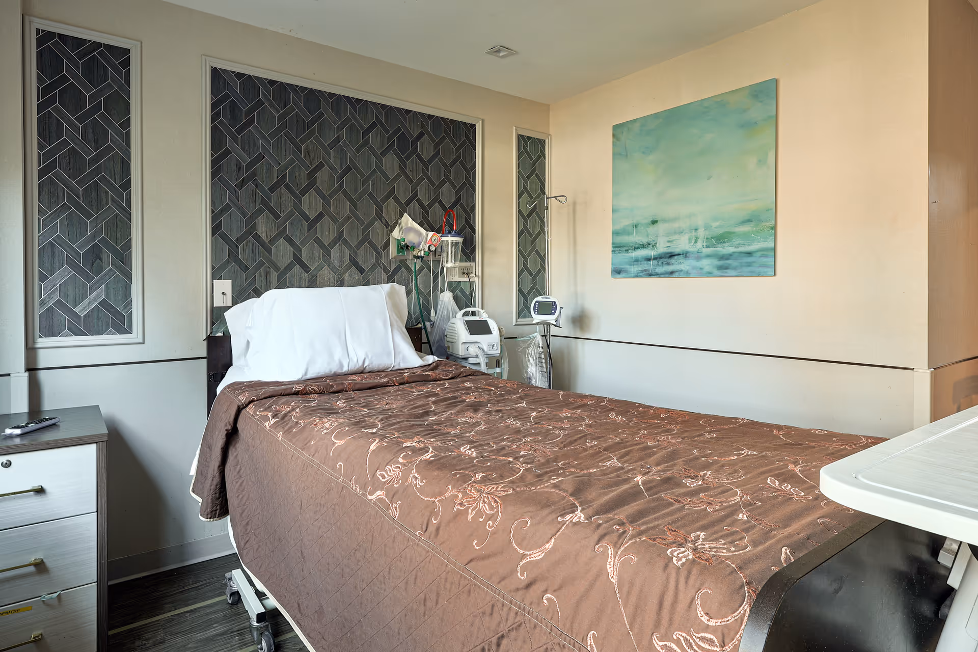 A nursing facility room with a single bed covered in a brown floral quilt. The bed has a white pillow and is positioned against a wall with decorative panels. Medical equipment is visible next to the bed, and a bedside table with drawers is on the left. A green and blue abstract painting hangs on the wall to the right.