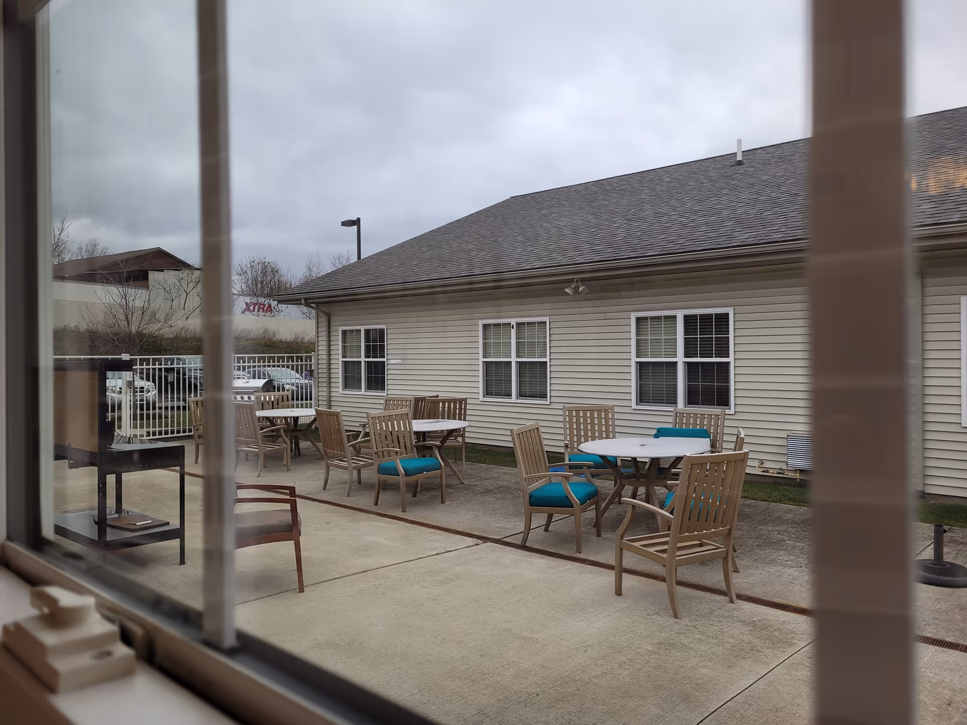 View through a window of an outdoor patio with round tables and wooden chairs beside a single-story building.