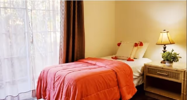 A cozy bedroom with a single bed covered in a coral-colored comforter and a decorative pillow with red tassels. Next to the bed is a wooden nightstand with a table lamp and a small potted plant. Sheer white curtains and brown drapes cover a large window, allowing natural light to brighten the room.
