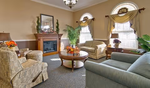 A cozy living room with a fireplace, two arched windows with yellow curtains, a round wooden coffee table with a floral centerpiece, two armchairs, a loveseat, and a sofa arranged around the table. There are plants and lamps on side tables, and a chandelier hanging from the ceiling.