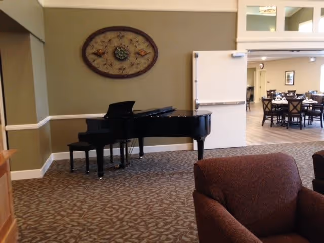 Interior view of a common area in an assisted living facility featuring a black grand piano with a matching bench against a green wall. Above the piano is a decorative oval wall hanging. In the foreground, there is a brown upholstered armchair. Through an open doorway, a dining area with tables and chairs is visible.