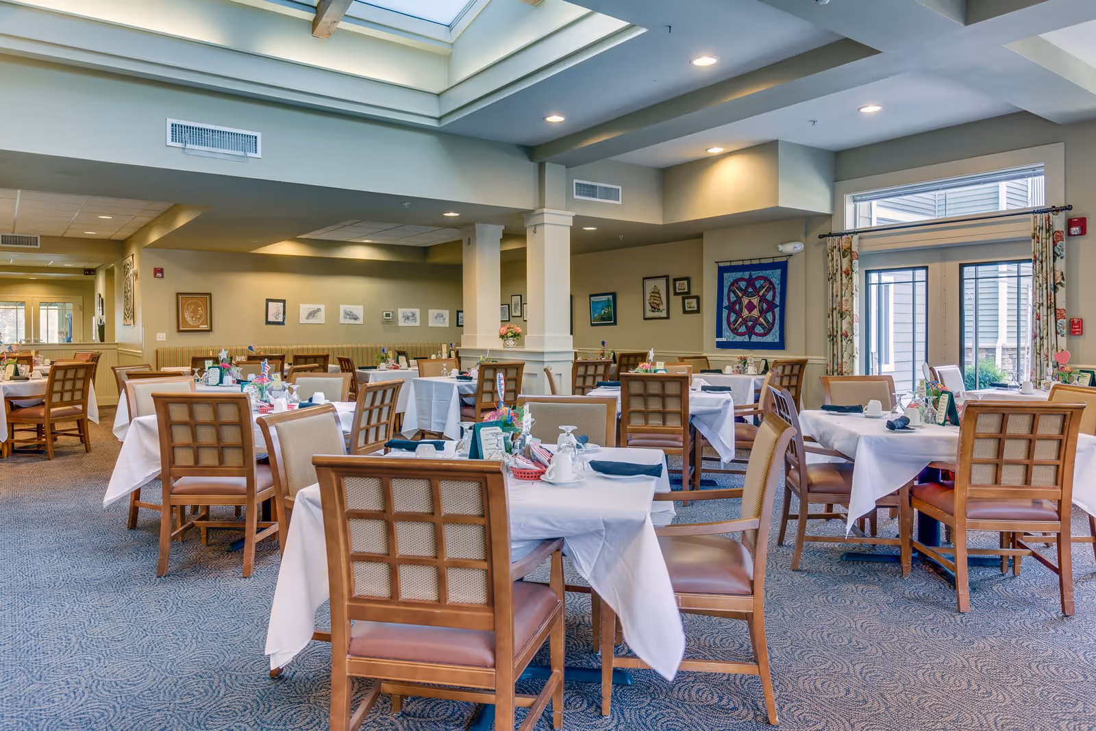 A spacious dining room with multiple tables covered in white tablecloths, each set with napkins, glasses, and small floral centerpieces. The room features wooden chairs with cushioned seats, carpeted floors, and soft lighting from ceiling fixtures. Large windows and a skylight provide natural light, and framed artwork decorates the walls.