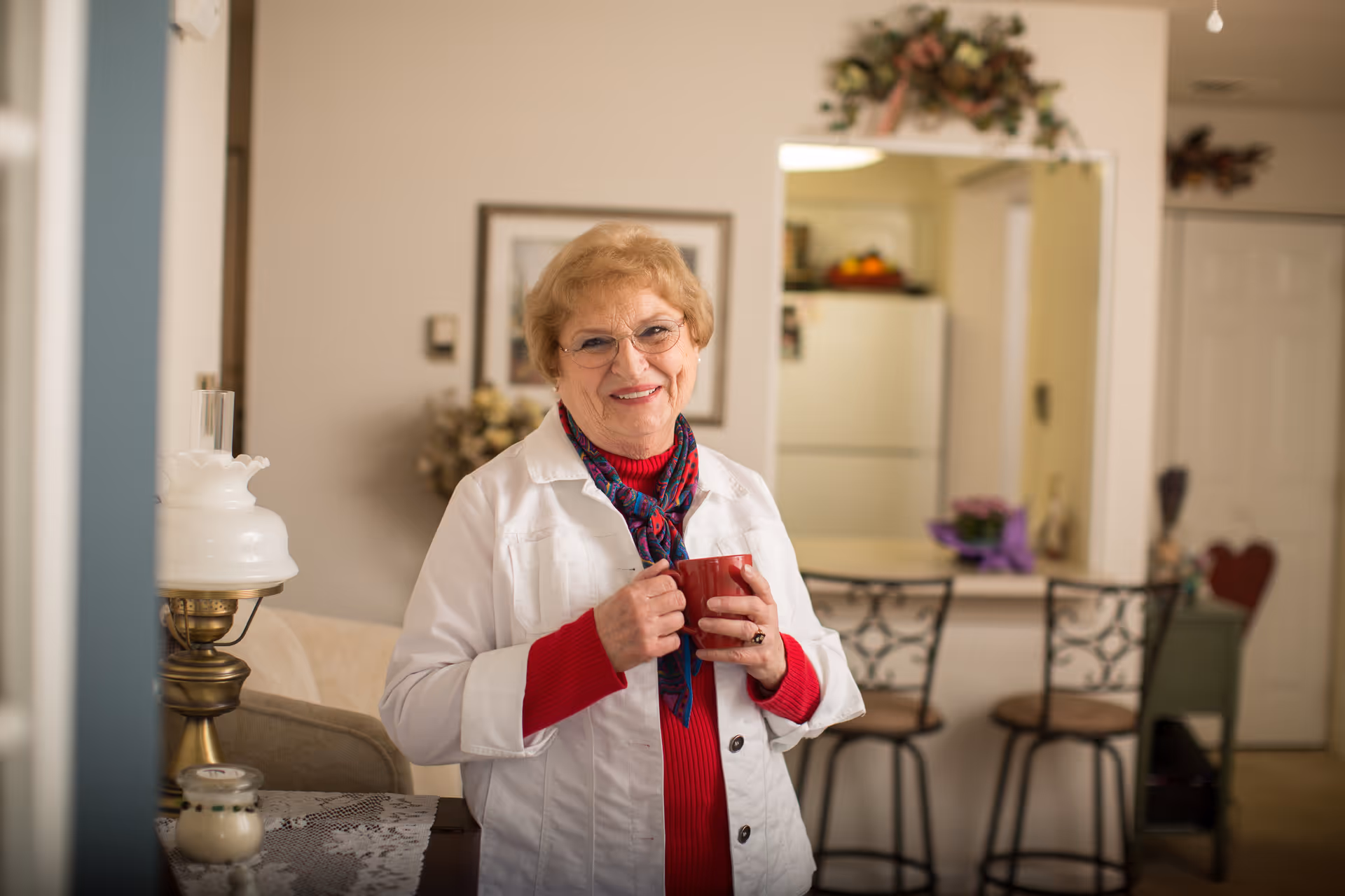 An elderly woman with glasses, wearing a white jacket and red sweater, stands indoors holding a red mug and smiling. Behind her is a kitchen area with a refrigerator, two bar stools, and a counter decorated with flowers.
