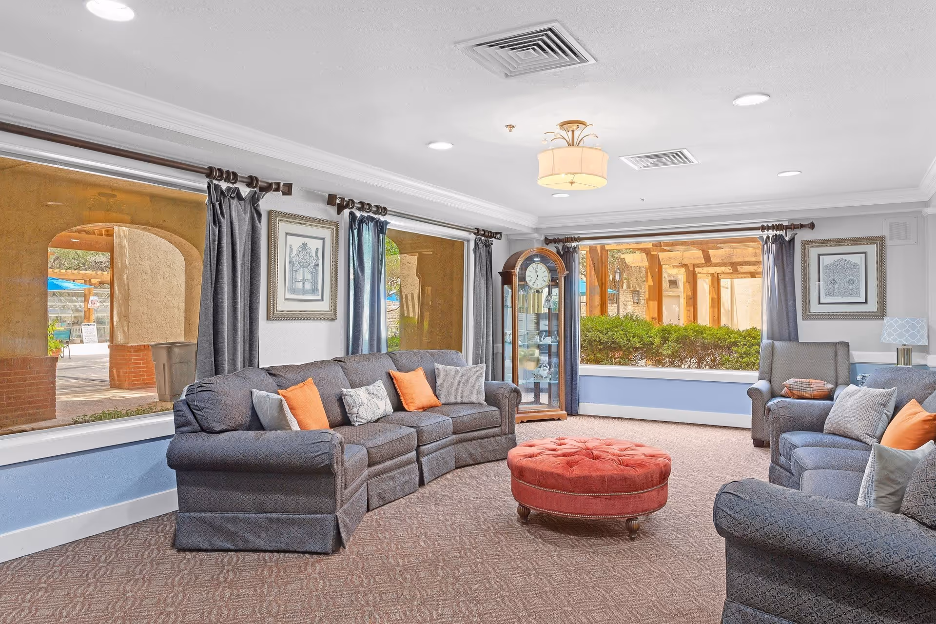 A bright and spacious living room with large windows showing an outdoor area. The room features two gray sofas with orange and gray cushions, a round red ottoman in the center, a grandfather clock in the corner, framed artwork on the walls, and gray curtains on the windows.