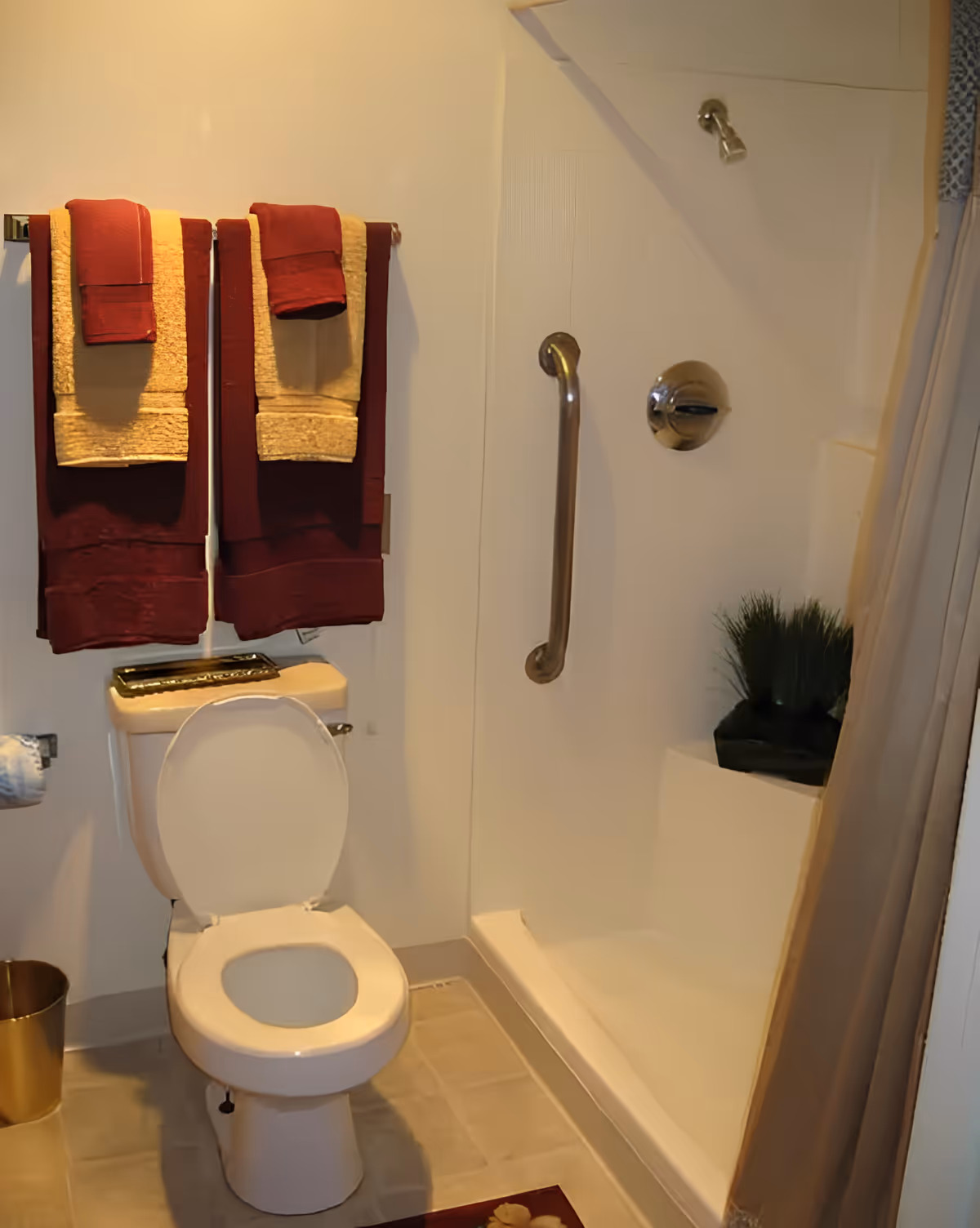 A bathroom featuring a white toilet with the lid open, a shower with a grab bar and showerhead, and a set of burgundy and beige towels hanging on a towel rack above the toilet. There is a small plant placed on the shower ledge and a beige shower curtain partially visible.