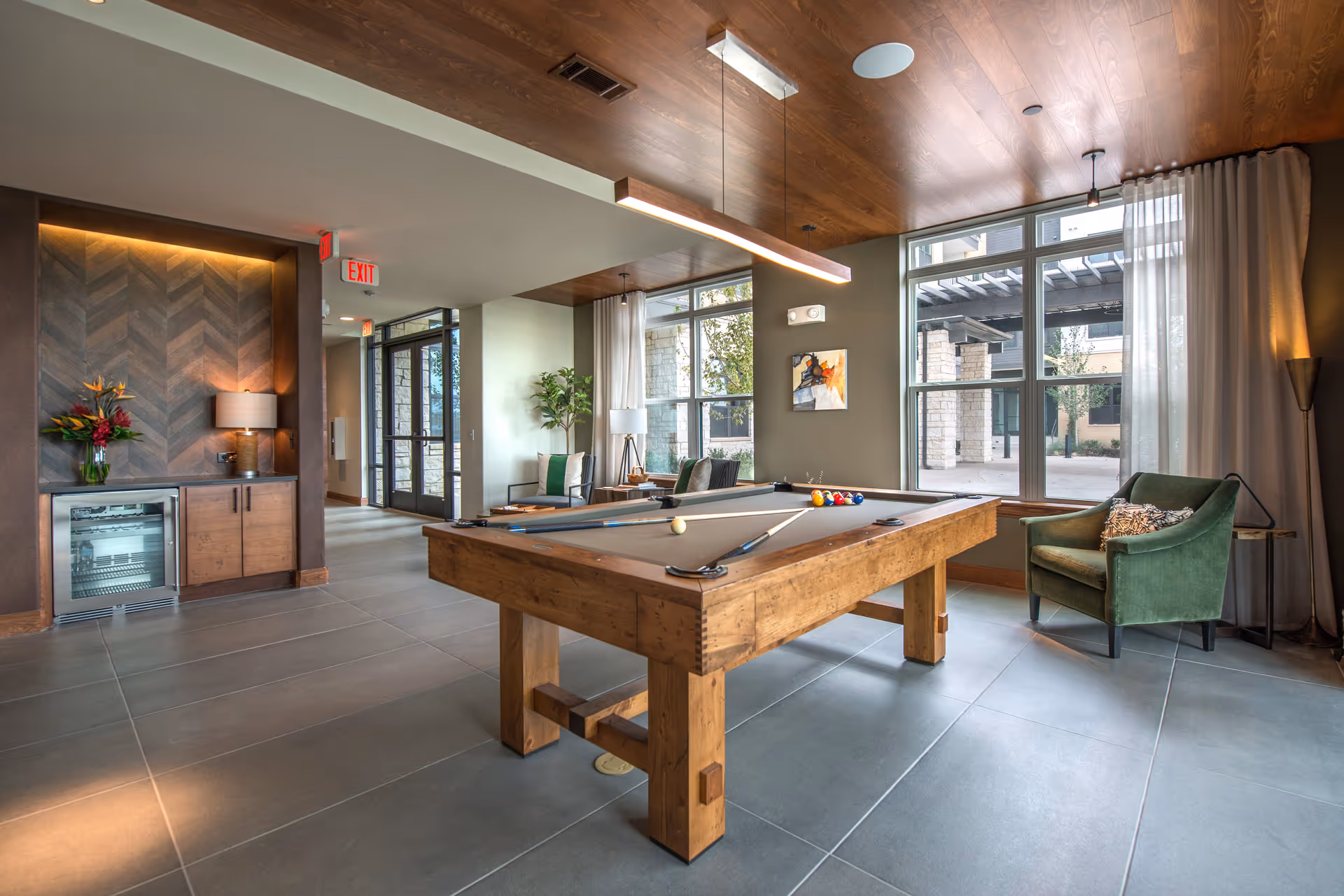 Bright communal game room with a wooden pool table, seating, large windows, and built-in cabinetry.