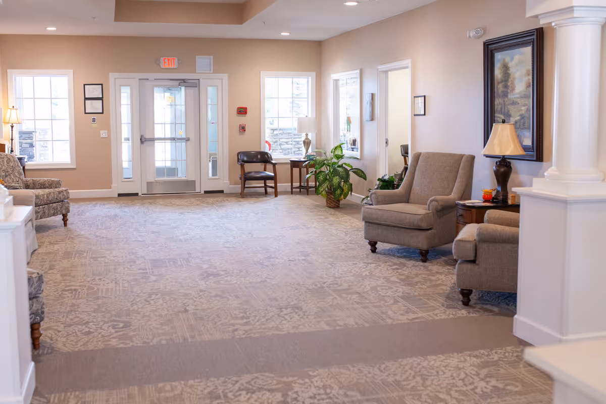 A bright and spacious assisted living facility common area with beige walls and patterned carpet. The room features several upholstered armchairs, a wooden side table with a lamp, a potted plant, framed artwork on the wall, and large windows letting in natural light. A glass exit door is visible at the far end of the room.