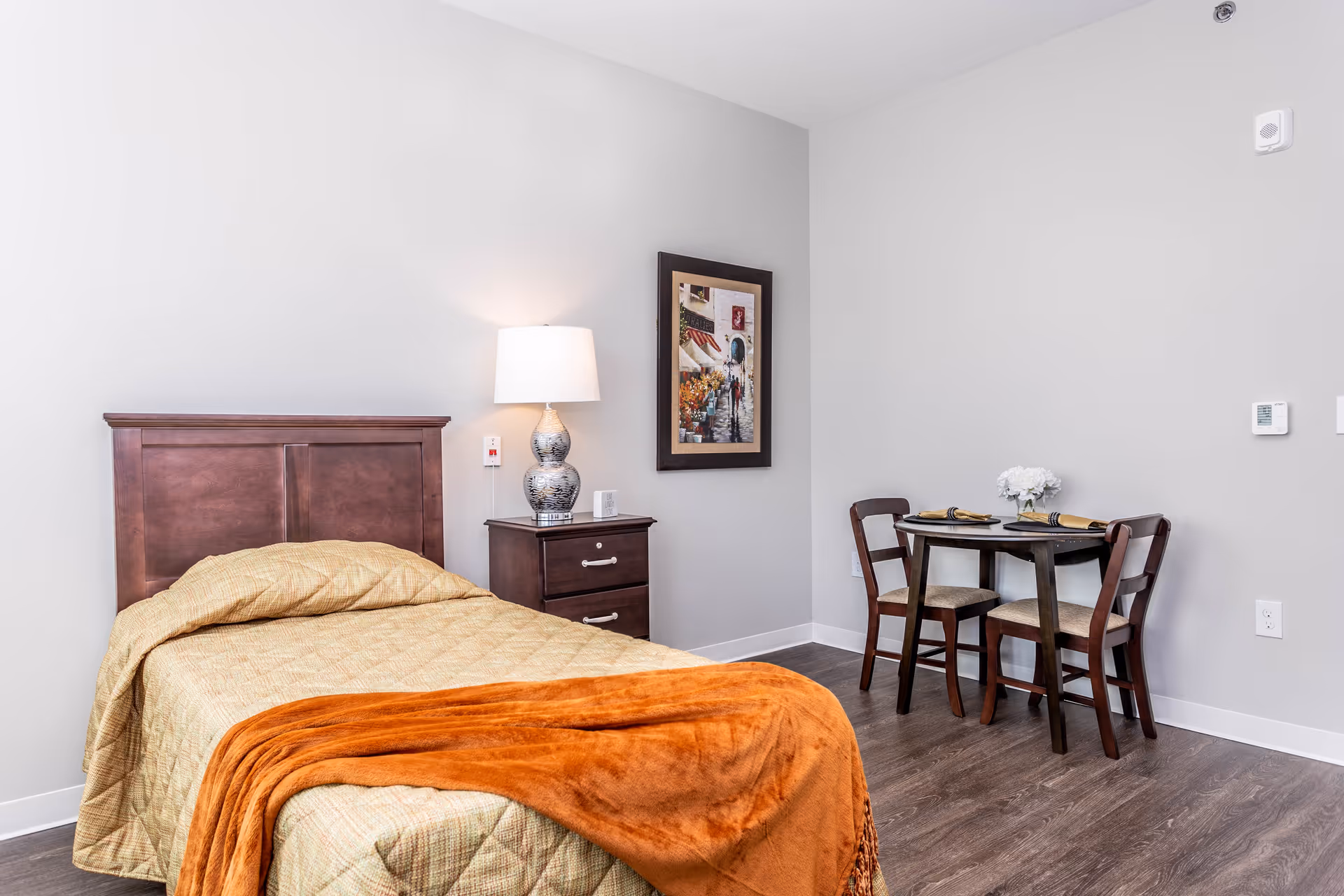 A senior living facility bedroom with a single bed covered in a beige quilt and an orange throw blanket. Next to the bed is a dark wooden nightstand with a decorative lamp and a small clock. On the wall above the nightstand is a framed picture. In the corner, there is a small round dining table set with two chairs, place settings, and a white flower centerpiece. The room has light gray walls and wood flooring.