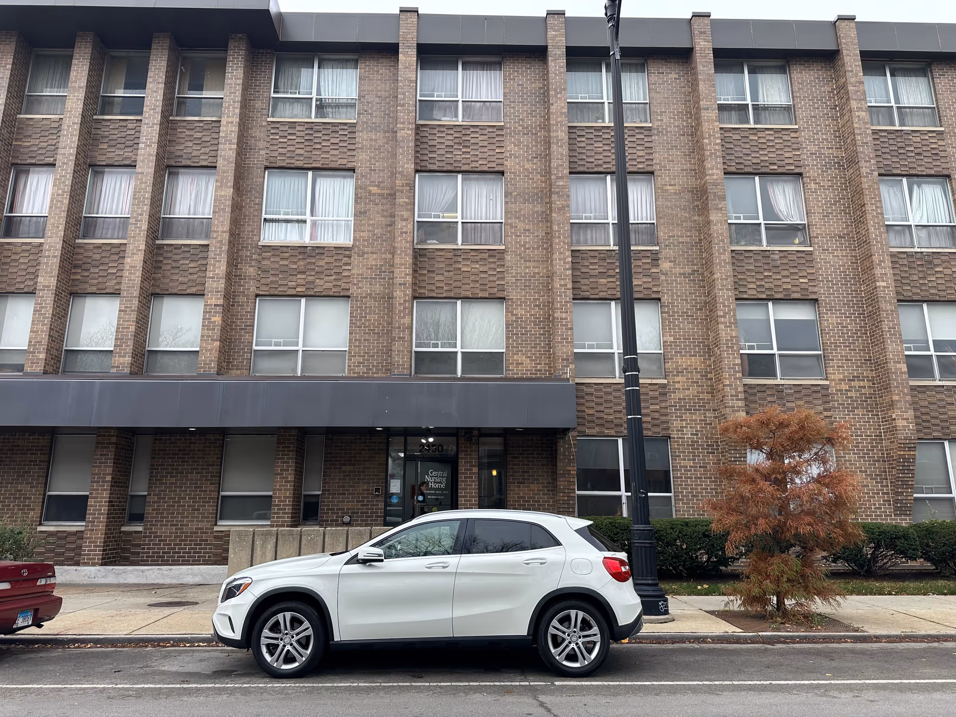 Exterior front view of a multi-story brick building with multiple windows, a black awning above the entrance, and a white car parked on the street in front. There is a small tree and a streetlamp near the sidewalk. The entrance door has a sign that reads 'Central Nursing Home'.