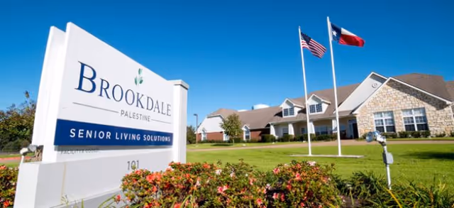 Exterior view of Brookdale Palestine senior living facility with a large sign in the foreground reading 'Brookdale Palestine Senior Living Solutions'. The building is a single-story structure with a stone and brick facade, surrounded by a well-maintained lawn and landscaping. Two flagpoles display the American and Texas flags against a clear blue sky.