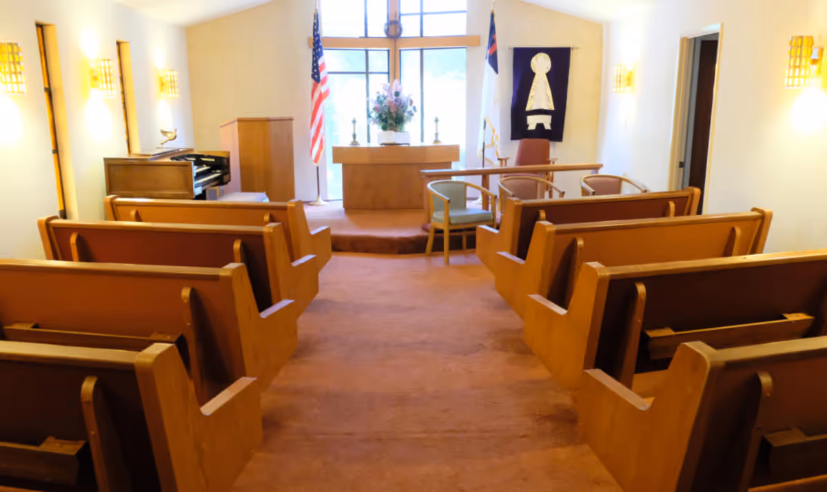 Interior view of a small chapel with wooden pews arranged in rows facing an altar. The altar has a floral arrangement and two candlesticks. There is an American flag and a Christian flag on either side of the altar. A piano is visible to the left, and there are wall-mounted lights on both sides of the room. The carpet is a muted orange color.