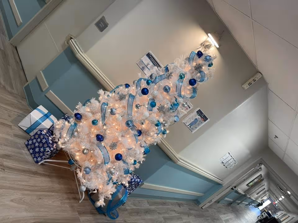 A white artificial Christmas tree decorated with blue, silver, and teal ornaments and ribbons, placed in a hallway with light blue and beige walls and wood-patterned flooring. Wrapped presents are placed under the tree. The hallway has ceiling lights and a few posters on the walls.