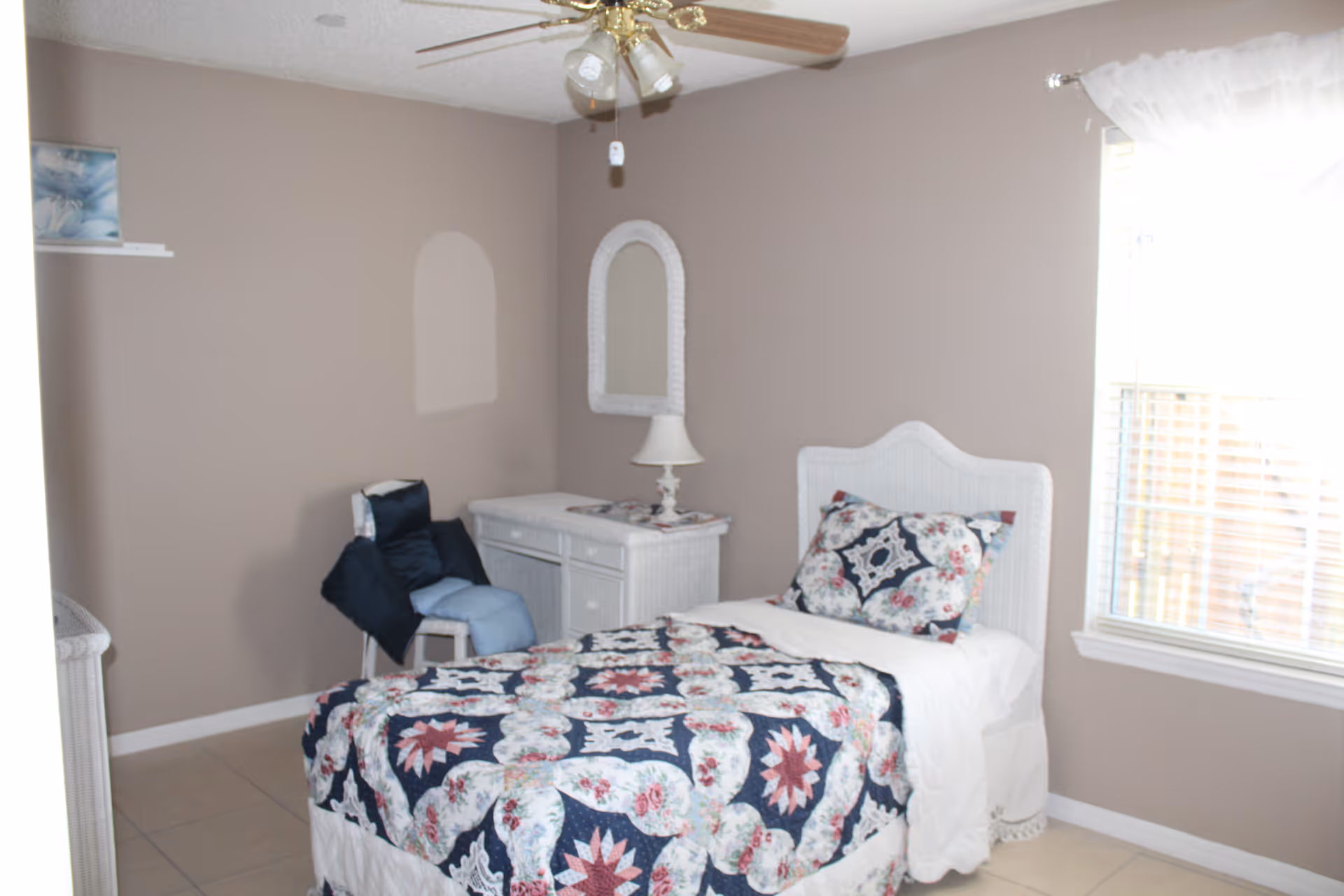 A small bedroom with a single bed covered in a floral and geometric patterned quilt in navy, white, and red. There is a white wicker headboard, a matching white wicker dresser with a lamp and a mirror above it, and a chair with cushions in the corner. A window with white curtains lets in natural light, and a ceiling fan with lights is mounted on the ceiling.