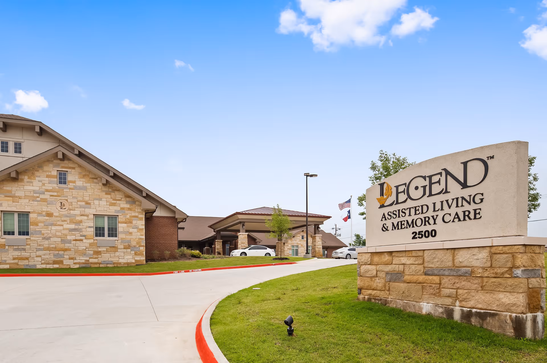 Exterior view of Legend Assisted Living & Memory Care facility showing a stone and brick building with a driveway and a large sign displaying the facility name and address 2500. The sky is clear with a few clouds.
