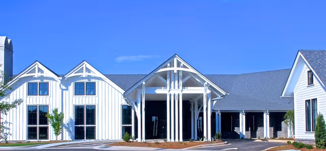 Front exterior view of a modern senior living facility building with white siding, large windows, and a covered entrance under a clear blue sky.