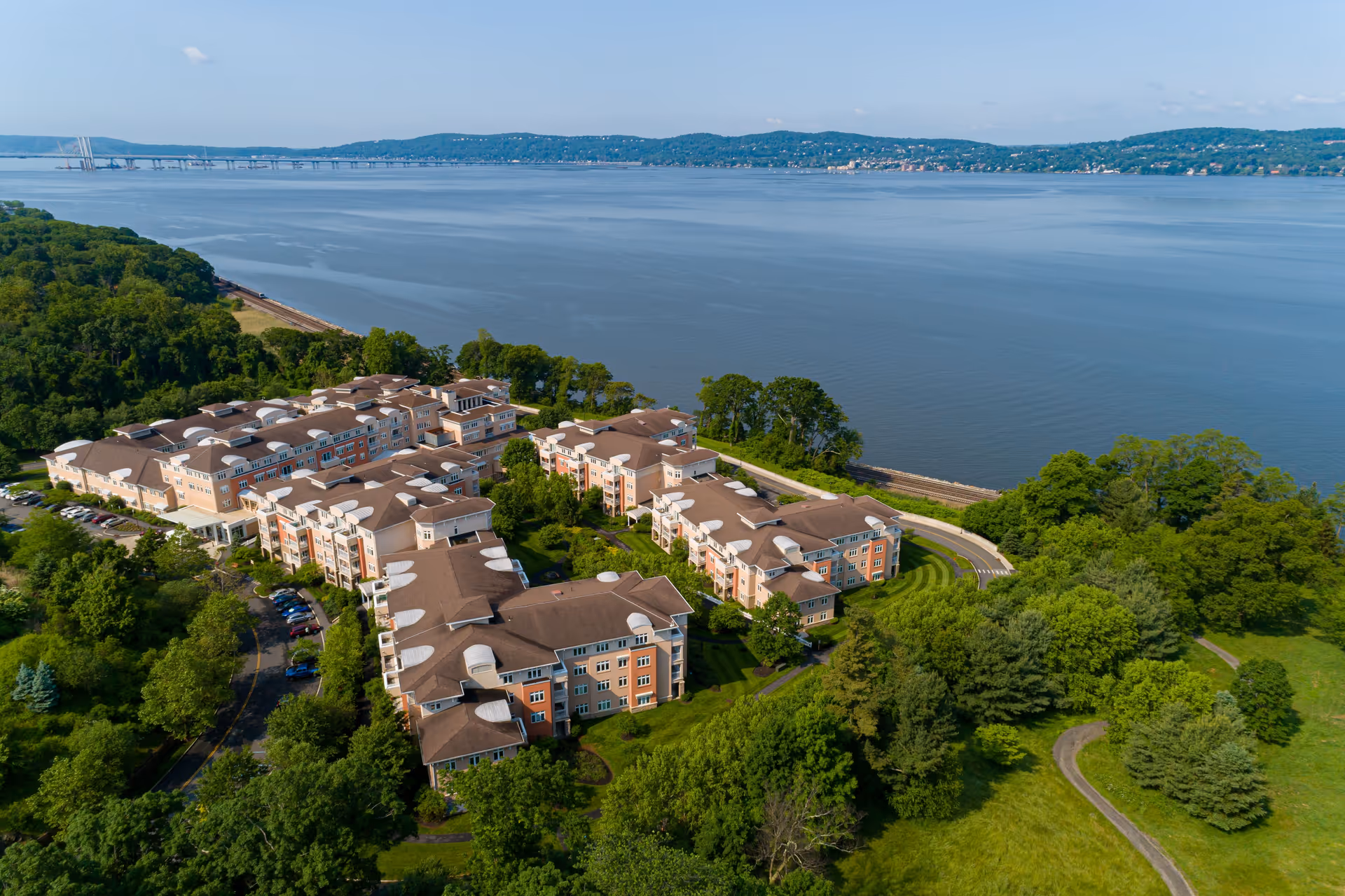 Aerial view of Kendal On Hudson senior living facility surrounded by lush green trees and lawns, located next to a large body of water with hills in the background and a bridge visible in the distance.