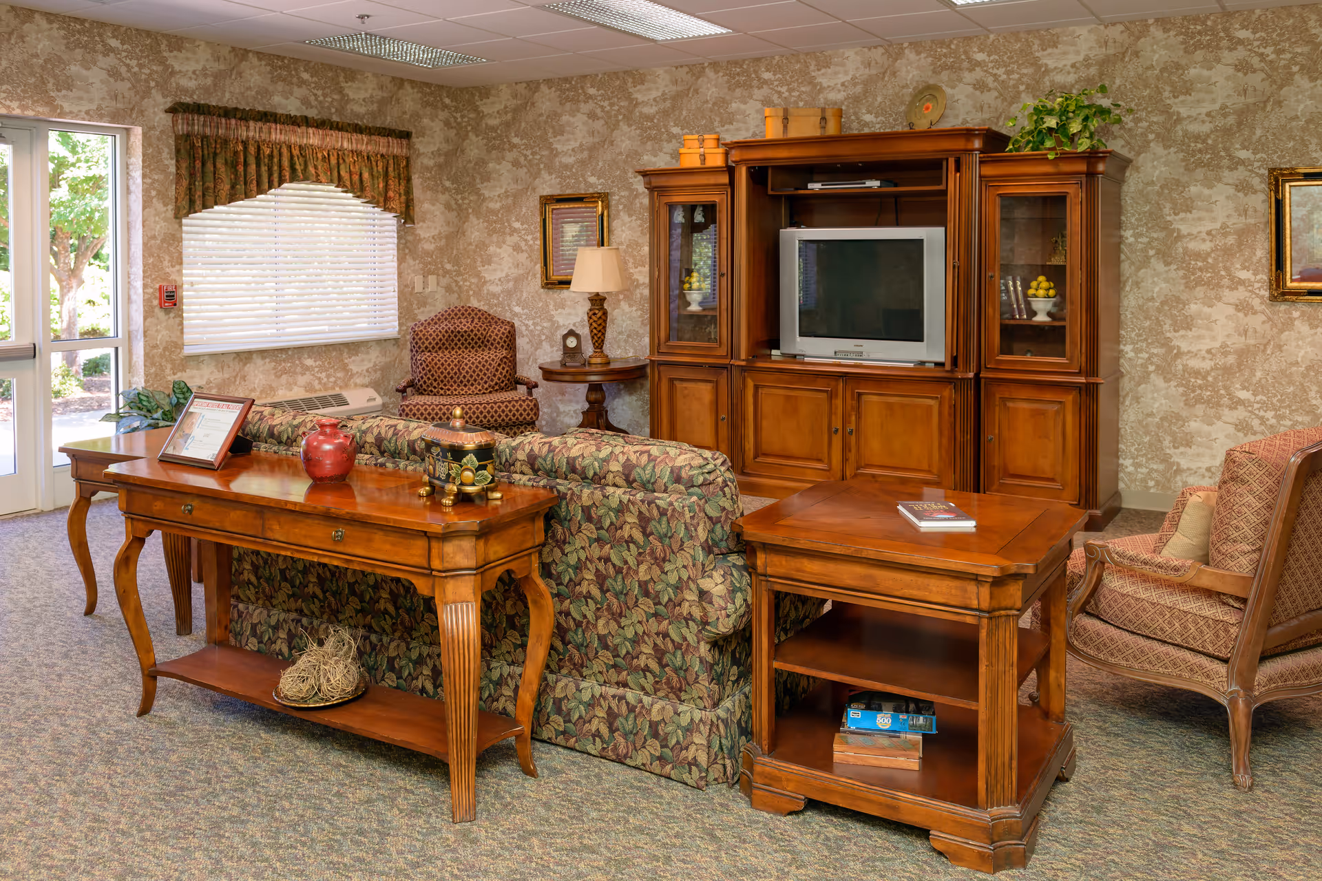 A cozy living room area in a senior living facility featuring a floral patterned sofa, wooden side and console tables, an armchair, and a wooden entertainment center with a television. The room has patterned wallpaper, a window with blinds and a valance, a table lamp, framed artwork, and a glass door leading outside.