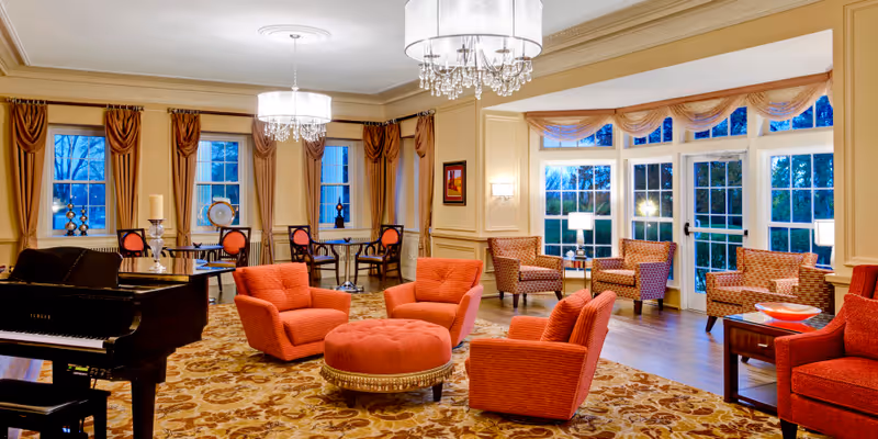 A spacious and elegant living room with large windows and glass doors letting in natural light. The room features a grand black piano, several red upholstered armchairs, a round ottoman, and patterned carpet. There are also small tables with lamps and decorative items, and chandeliers hanging from the ceiling.