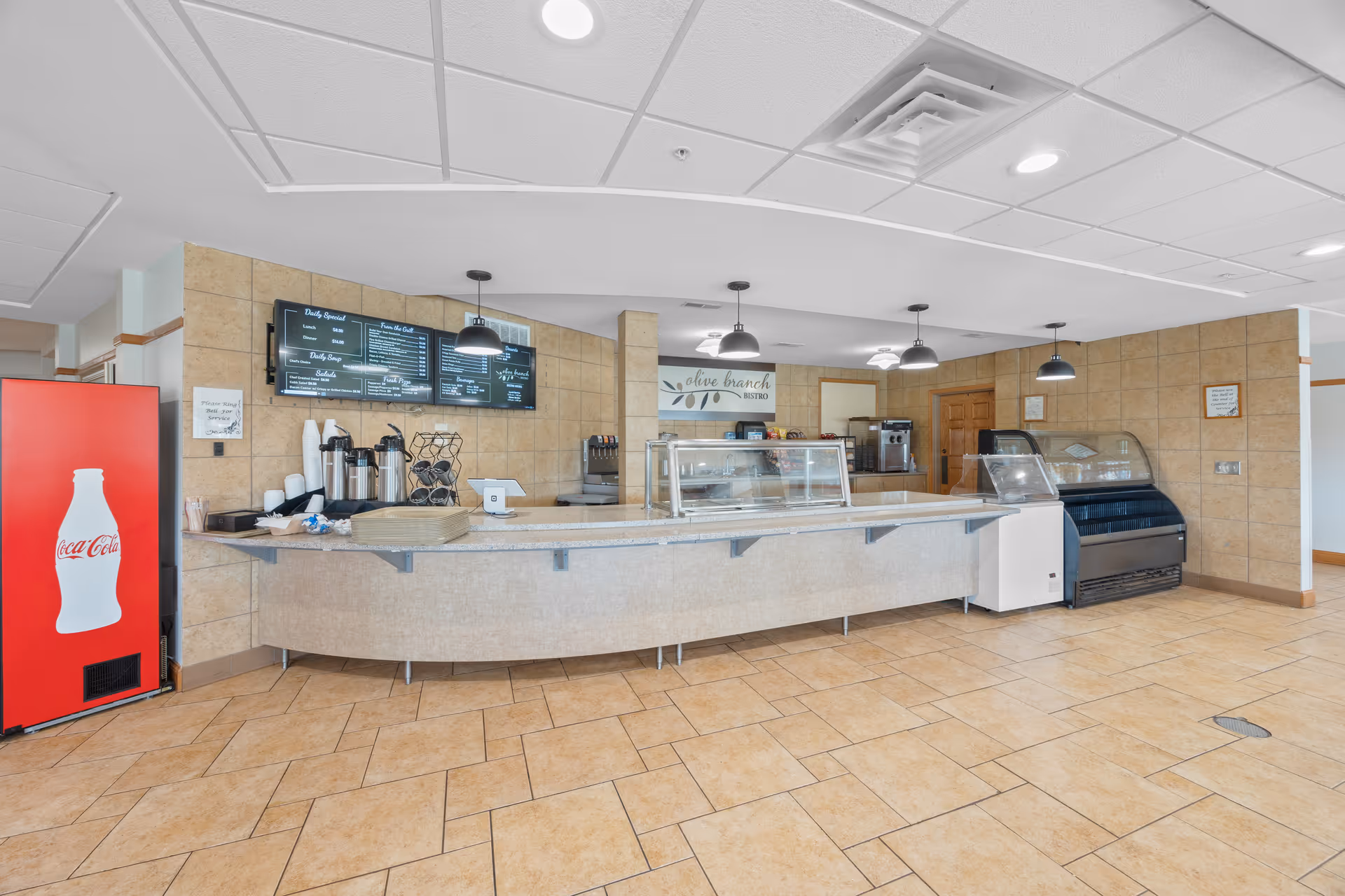 Cafeteria service counter with glass display cases, menu boards, beverage equipment, and a Coca-Cola vending machine.