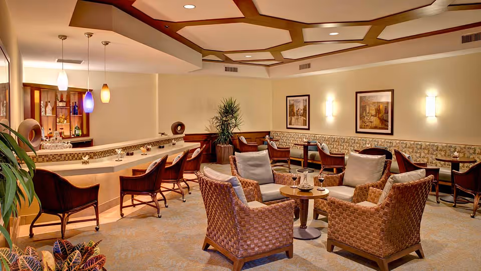 A cozy lounge area with wicker armchairs and small round tables, a bar counter with high chairs, pendant lights hanging from the ceiling, framed artwork on the walls, and a potted plant in the corner.
