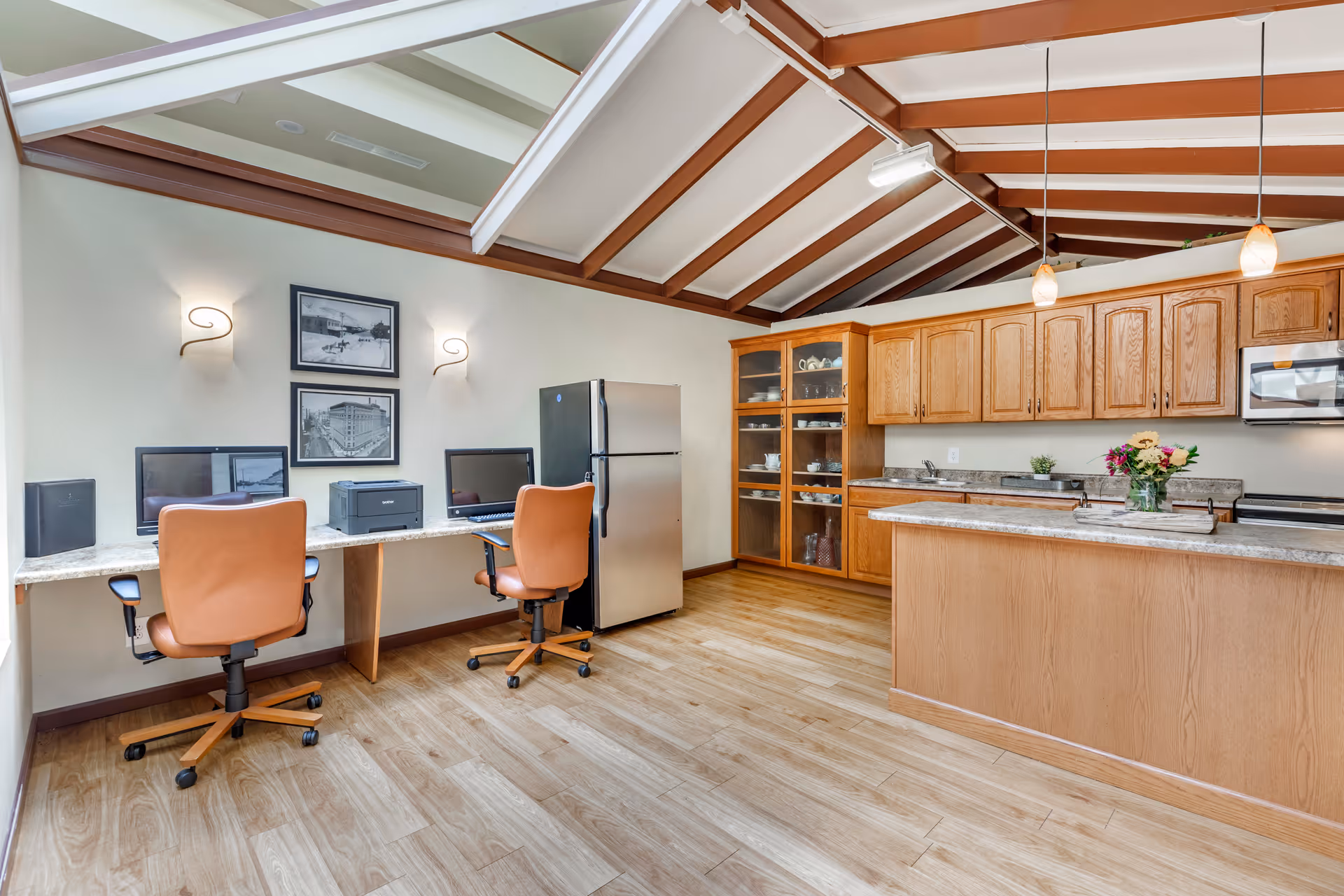 Interior view of a room with a kitchen area and a small workspace. The kitchen has wooden cabinets, a countertop with a vase of flowers, a stainless steel refrigerator, and a microwave. The workspace includes two desks with computers, a printer, and two brown office chairs. The ceiling has exposed beams and pendant lights.