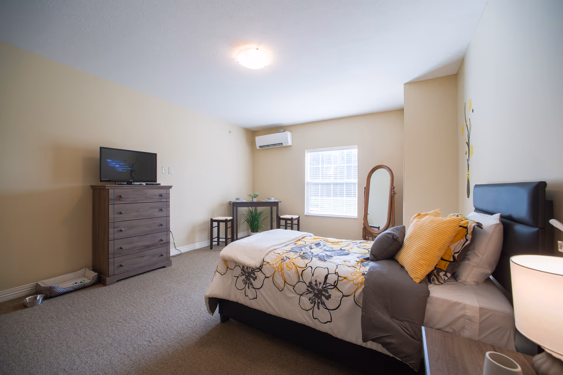 Bright furnished bedroom with a floral bed, dresser with TV, window and full-length mirror.