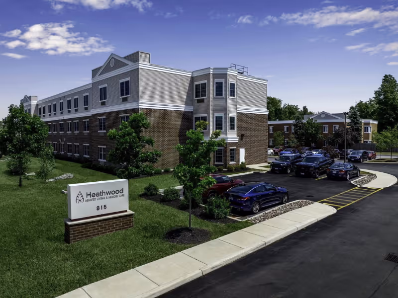 Front exterior of a multi-story brick and siding assisted living building with a parking lot and a sign reading Heathwood Assisted Living & Memory Care.