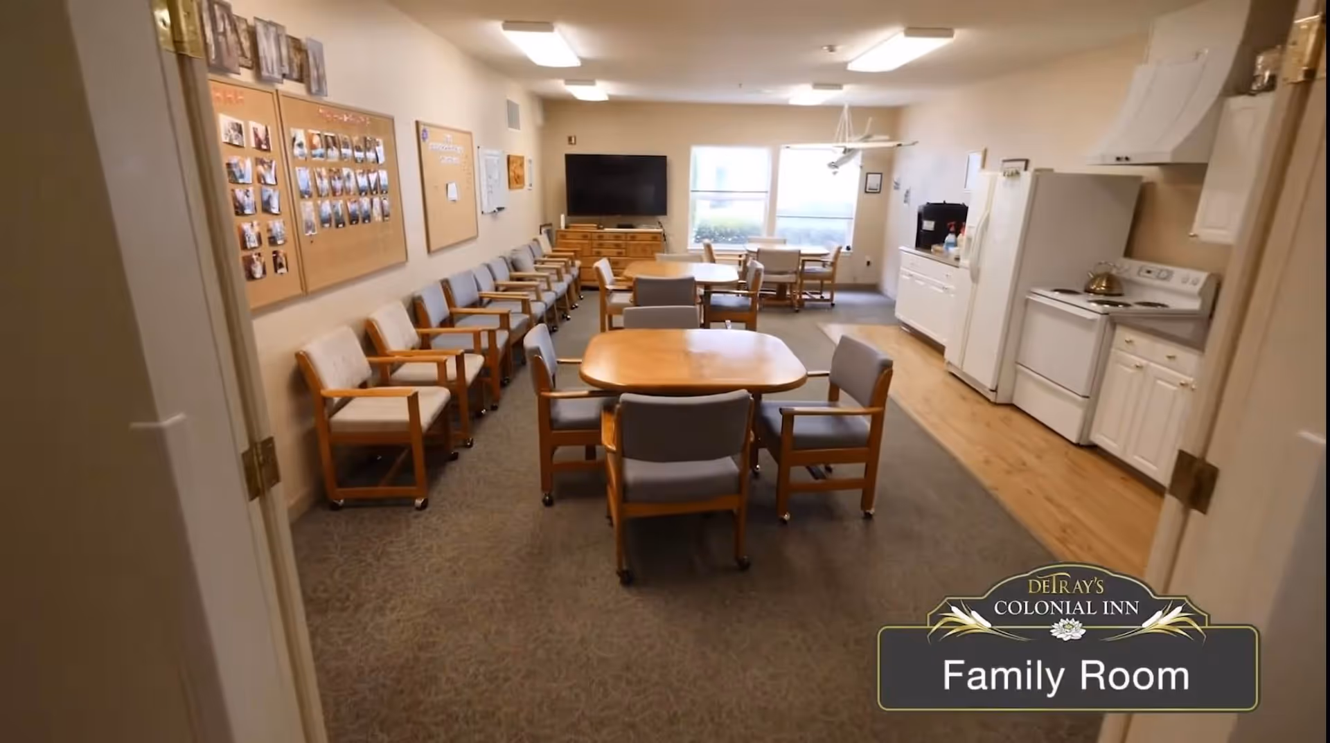 Interior view of a family room in DeTray's Colonial Inn Retirement Apartments featuring multiple wooden tables with cushioned chairs, a row of chairs lined up against the wall, a flat-screen TV mounted on a dresser, and a kitchenette area with a refrigerator, stove, and cabinets.