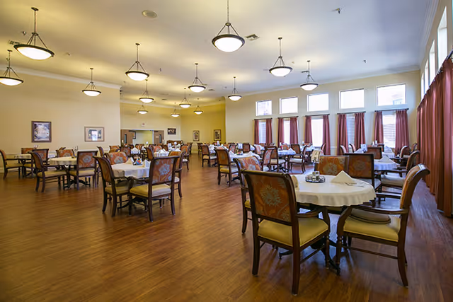 A spacious dining room with multiple round tables covered with white tablecloths, each surrounded by wooden chairs with patterned cushions. The room has wooden flooring, several hanging ceiling lights, large windows with red curtains, and framed pictures on the walls.