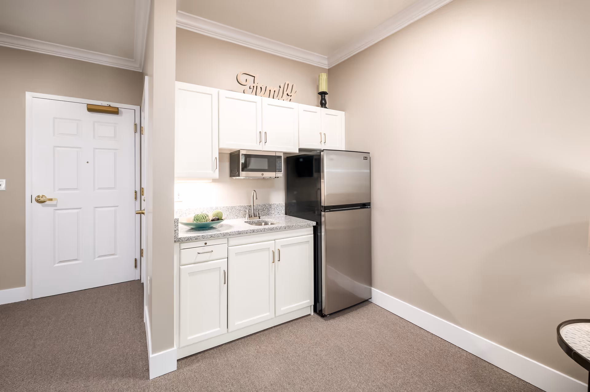 Small kitchenette area with white cabinets, a granite countertop, a stainless steel refrigerator, a microwave mounted above the sink, and a decorative bowl with green spheres on the counter. The walls are beige, and there is a white door to the left. A decorative word 'Family' is displayed above the cabinets.