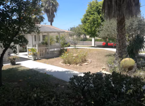 Outdoor view of a garden area at Acorn Oaks Manor I featuring a small white gazebo with a roof, surrounded by trees including palm trees and various plants. There is a concrete pathway leading to the gazebo and a red car visible in the background near a white fence.