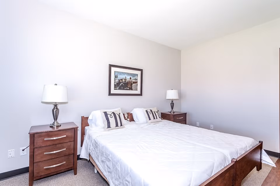 A bedroom with a large bed covered in a white quilt and two pillows labeled 'Mr' and 'Mrs'. There are two wooden nightstands on either side of the bed, each with a silver lamp. A framed picture hangs on the wall above the bed. The walls are light-colored and the floor is carpeted.