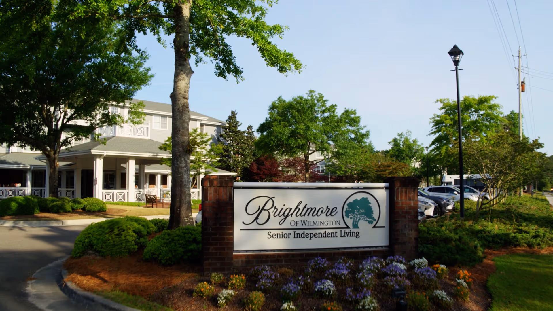 Exterior view of Brightmore of Wilmington Senior Independent Living facility with a large sign in the foreground surrounded by flowers and greenery, a tree, and a building with a porch in the background under a clear sky.