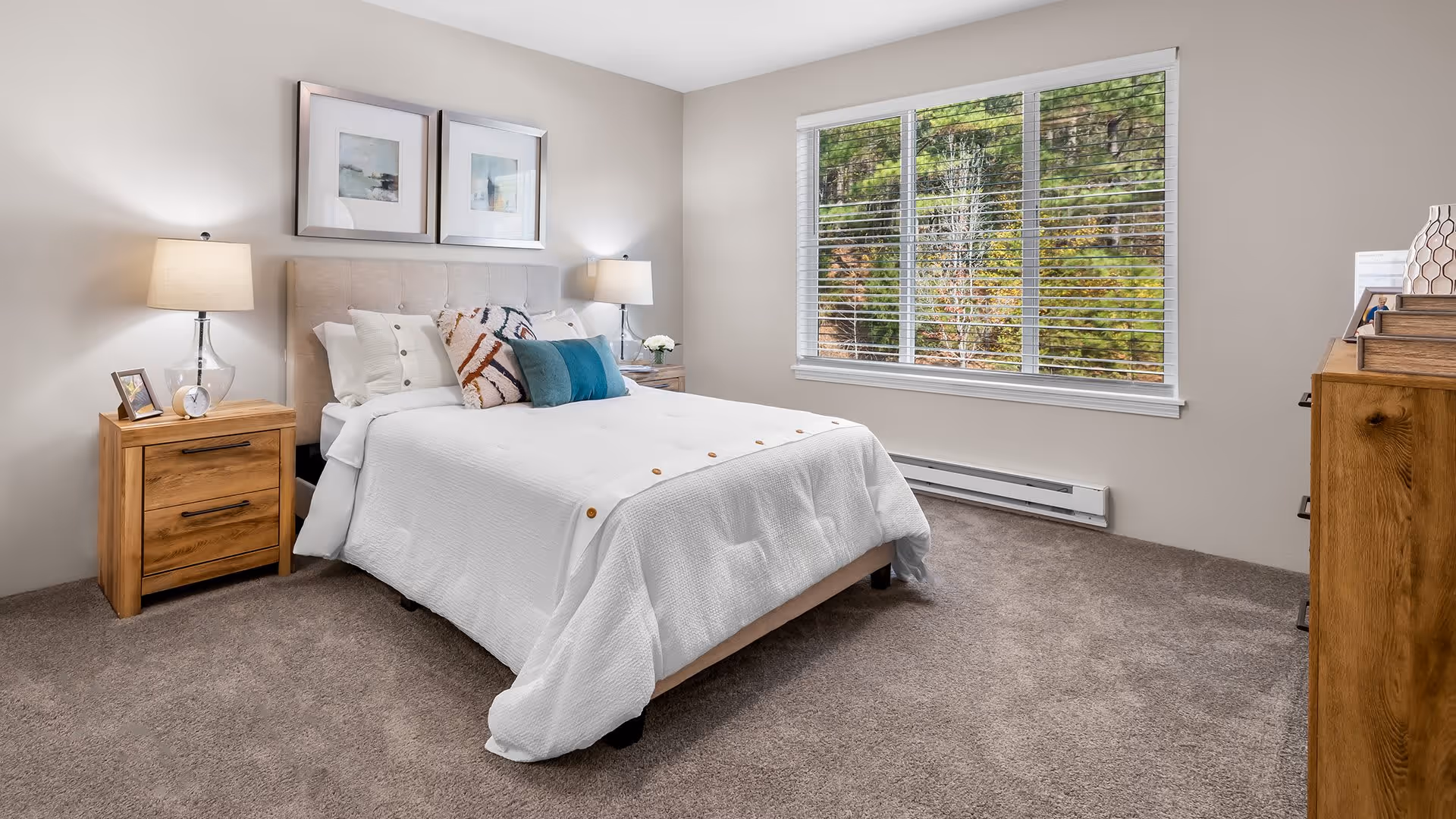 A neatly made bed with white bedding and decorative pillows in a bedroom. There are two wooden nightstands on either side of the bed, each with a lamp and small decorative items. Above the bed are two framed pictures. A large window with white blinds shows greenery outside. The room has beige walls and carpeted floor.