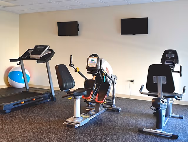 A fitness room with exercise equipment including a treadmill, a recumbent bike, and a rowing machine. Two wall-mounted televisions are visible on a beige wall, and a large blue and white exercise ball is placed near the treadmill.
