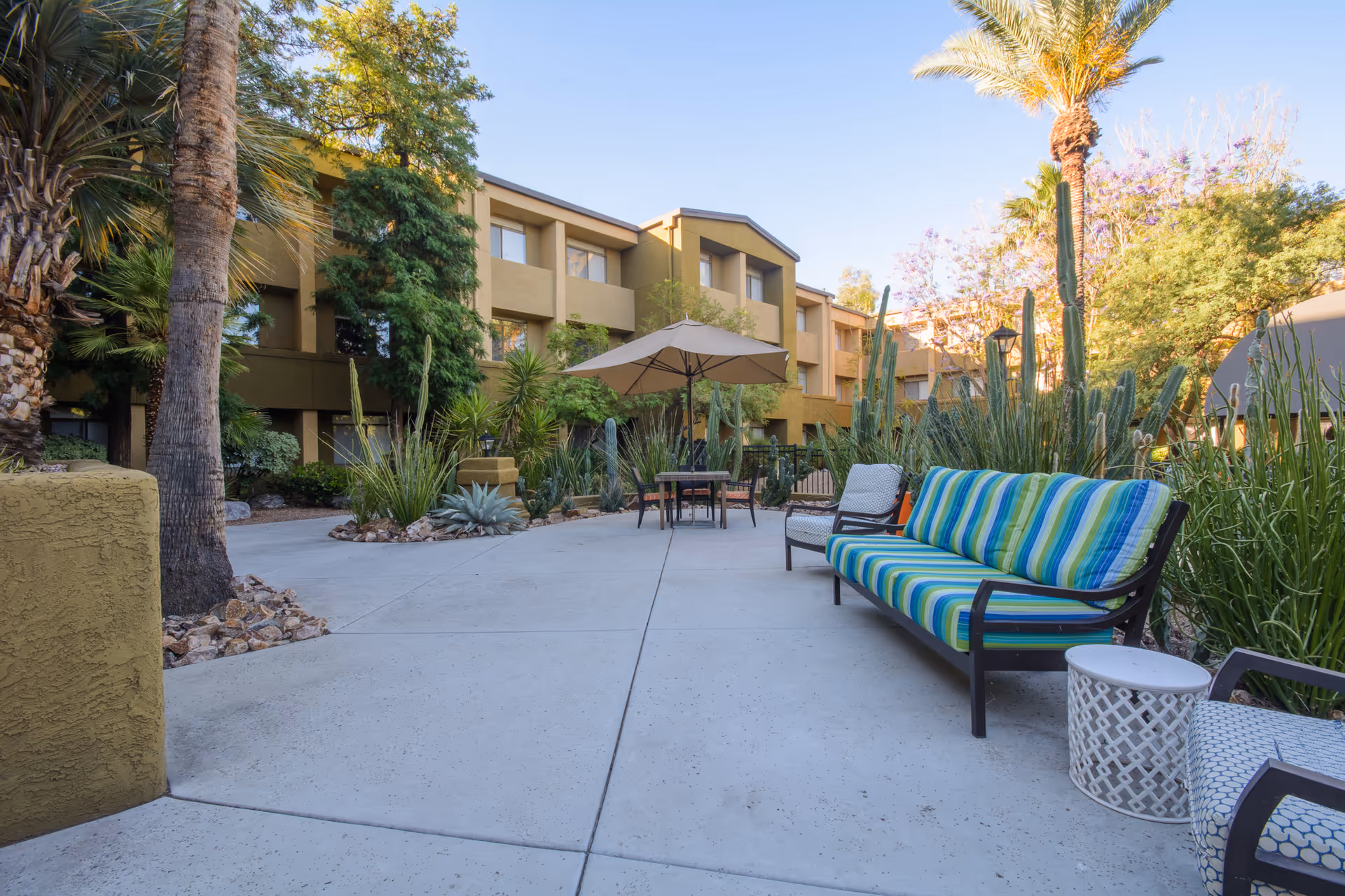 Outdoor courtyard with striped cushioned benches, patio tables and umbrella surrounded by palm trees, succulents and a multi-story building.