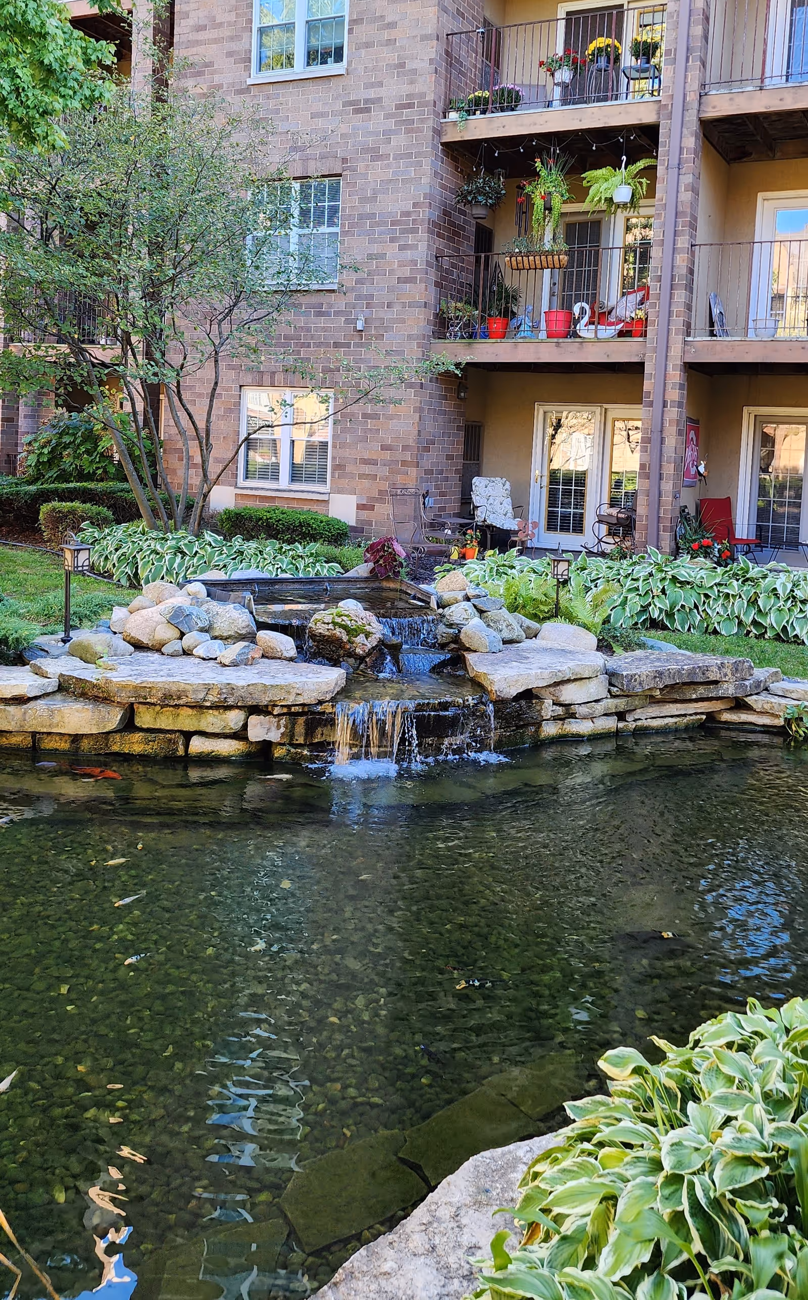 A serene outdoor garden area at The Commons Corewell Health - Oak Terrace Dearborn featuring a small cascading waterfall over rocks flowing into a clear pond surrounded by green plants. In the background, there is a brick building with balconies decorated with potted plants and outdoor furniture.