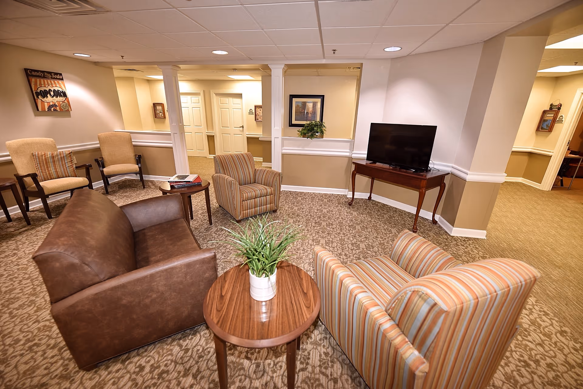 A cozy senior living common area with a brown leather loveseat, two striped armchairs, and two beige armchairs arranged around small wooden tables. A flat-screen TV sits on a wooden table against the wall. The room has beige walls, carpeted floors, and recessed ceiling lights. There is a decorative plant on one of the tables and framed artwork on the walls.