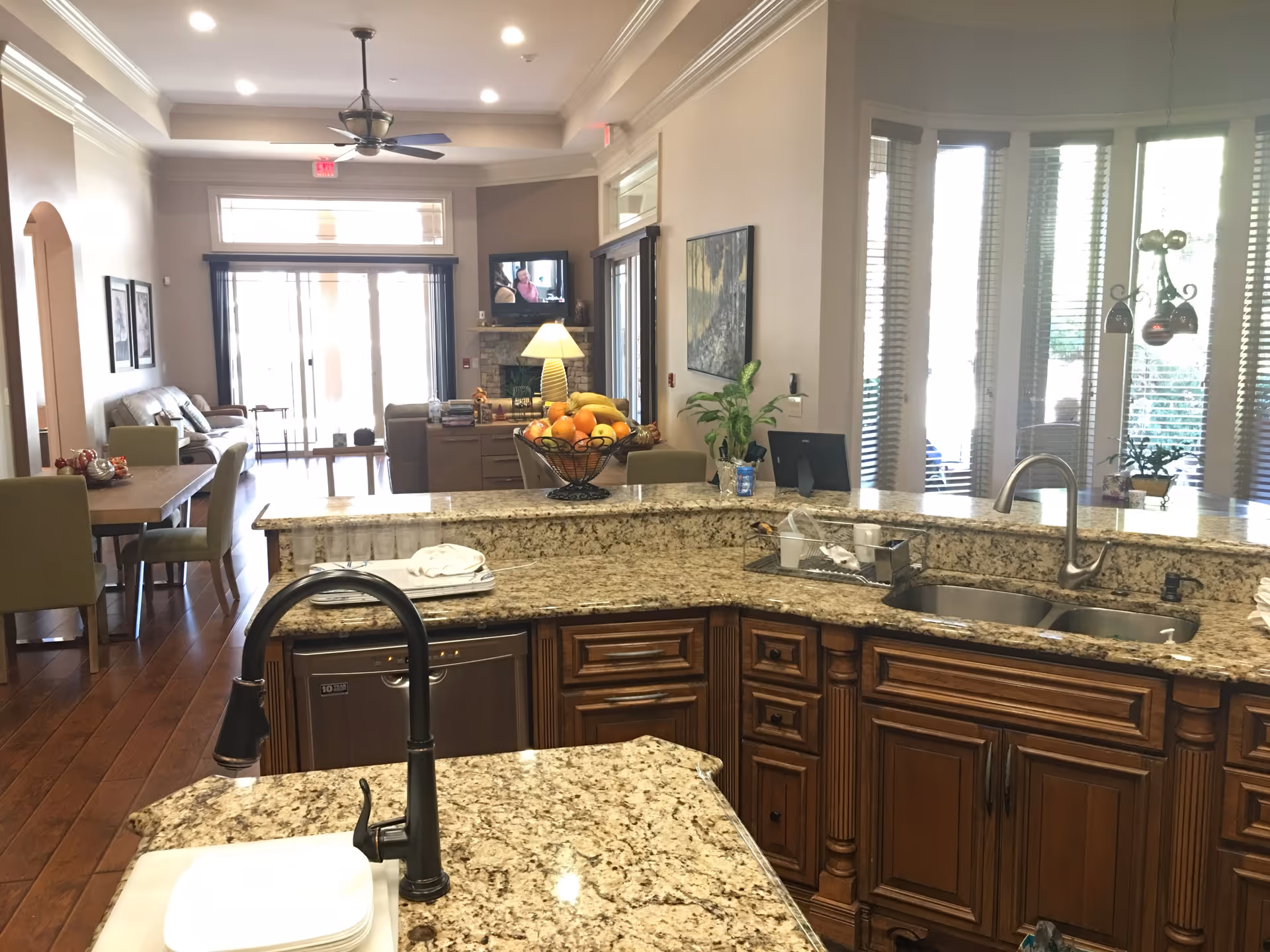 View of a spacious kitchen with granite countertops and wooden cabinets. The kitchen features a double sink, a black faucet, and a dishwasher. In the background, there is a dining area with a table and chairs, a living room with sofas, a ceiling fan, and a TV mounted above a fireplace. Large windows with blinds allow natural light to fill the space.