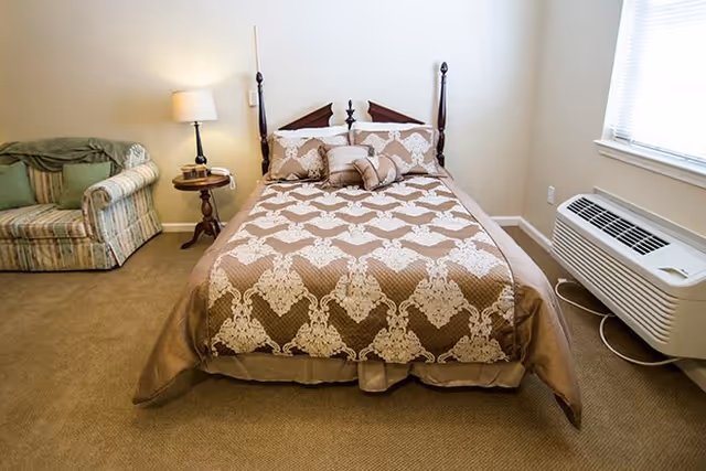 A bedroom with a bed covered in a beige and white patterned bedspread, two matching pillows, and a small decorative pillow. To the left of the bed is a wooden side table with a lamp and a phone, and a striped armchair with a green blanket draped over the back. To the right is a window with blinds and an air conditioning unit below it. The room has beige carpet and light-colored walls.