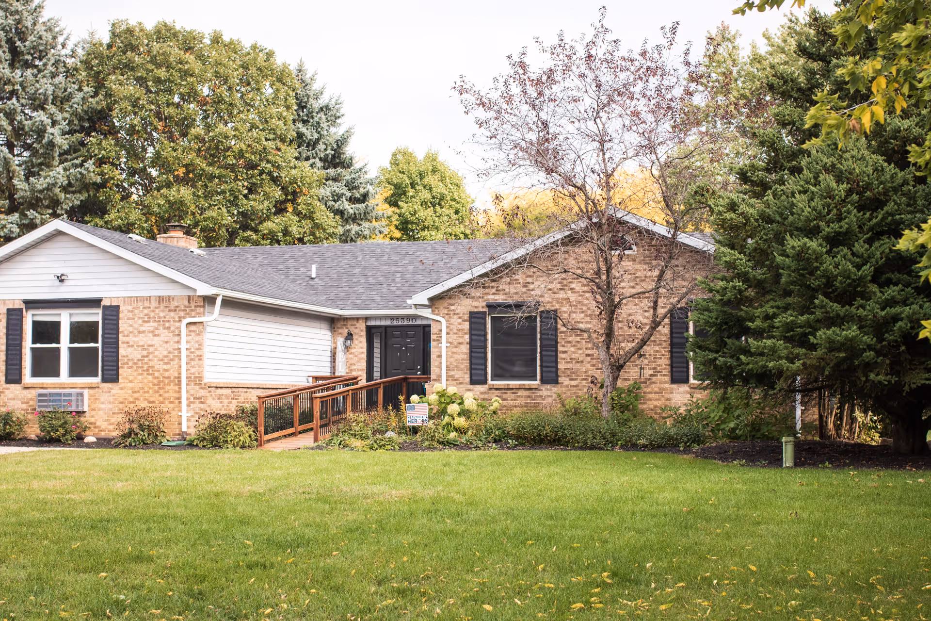 Front exterior view of a single-story brick building with a dark shingled roof, black window shutters, and a wooden ramp leading to a black front door. The building is surrounded by green grass, bushes, and trees with some autumn foliage.