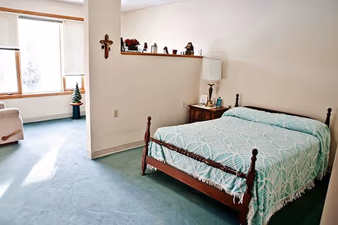 A bedroom with a wooden bed frame covered with a light blue patterned bedspread. Next to the bed is a wooden nightstand with a lamp and some small items. The room has light-colored walls and green carpet. There is a small alcove with a window and a small decorative Christmas tree on a stand. A cross and several small decorative items are displayed on a ledge above the nightstand.