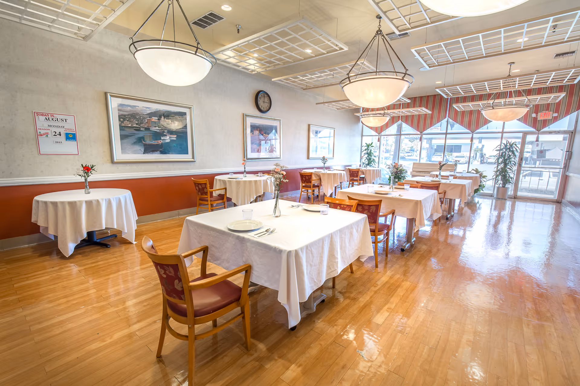 A bright dining room with several tables covered in white tablecloths, each set with plates, glasses, and silverware. Wooden chairs with red cushions surround the tables. The room has large windows with striped valances, letting in natural light. There are framed paintings on the walls, a clock, and hanging light fixtures above the tables. The floor is polished wood, and there are small flower vases on each table.