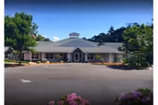 Single-story assisted living building with a central entrance, circular driveway and landscaped trees and shrubs under a blue sky.