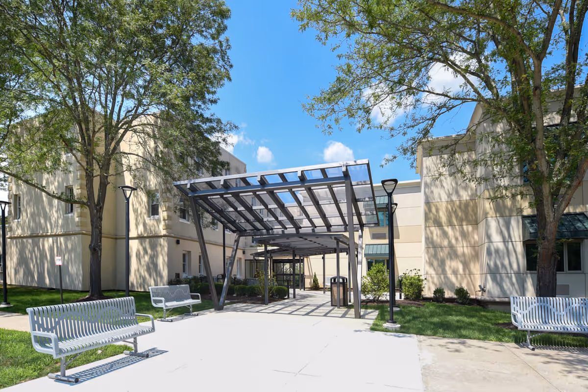 Entrance courtyard of a senior living facility with a covered metal walkway, benches, trees, and beige building facades.