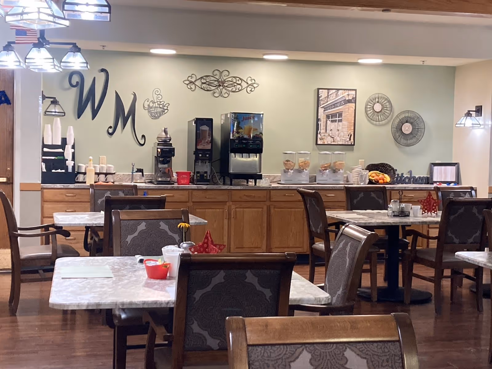 Dining room with tables and chairs and a self-serve beverage and cereal station along a back counter under wall decor reading 'WM'.