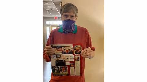 An elderly person wearing a purple face mask and a red shirt stands indoors holding a colorful printed page with various images and text. The background shows a hallway with a door and windows.