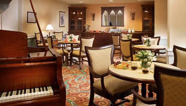 A cozy dining area with round tables and upholstered chairs arranged on a patterned carpet. A grand piano is visible on the left side of the room. The back wall features a wooden cabinet with decorative items and a mirror above it. The room is warmly lit with wall sconces and a table lamp.